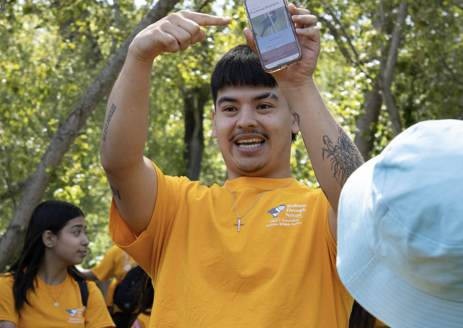 Person in a yellow T-shirt holding up a smartphone in a wooded area.